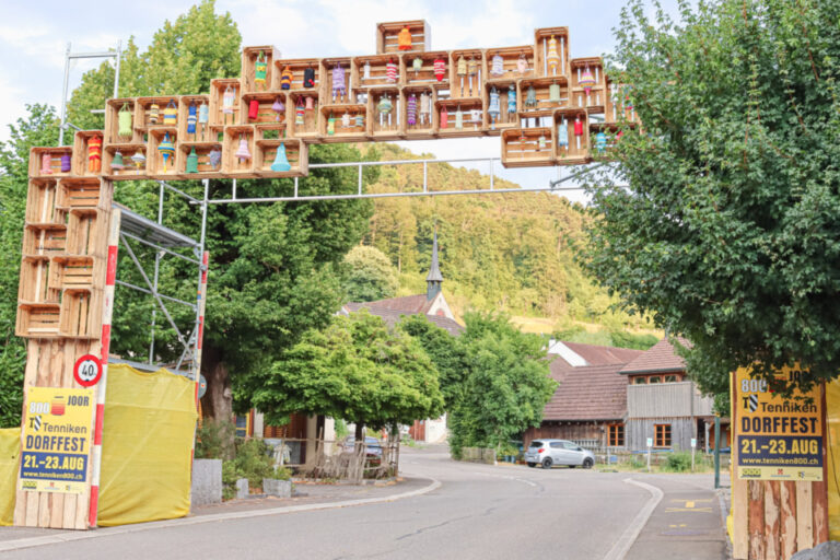 Ein Torbogen aus bunt bemalten Holzkisten überspannt eine Dorfstrasse vor grünen Hügeln.
