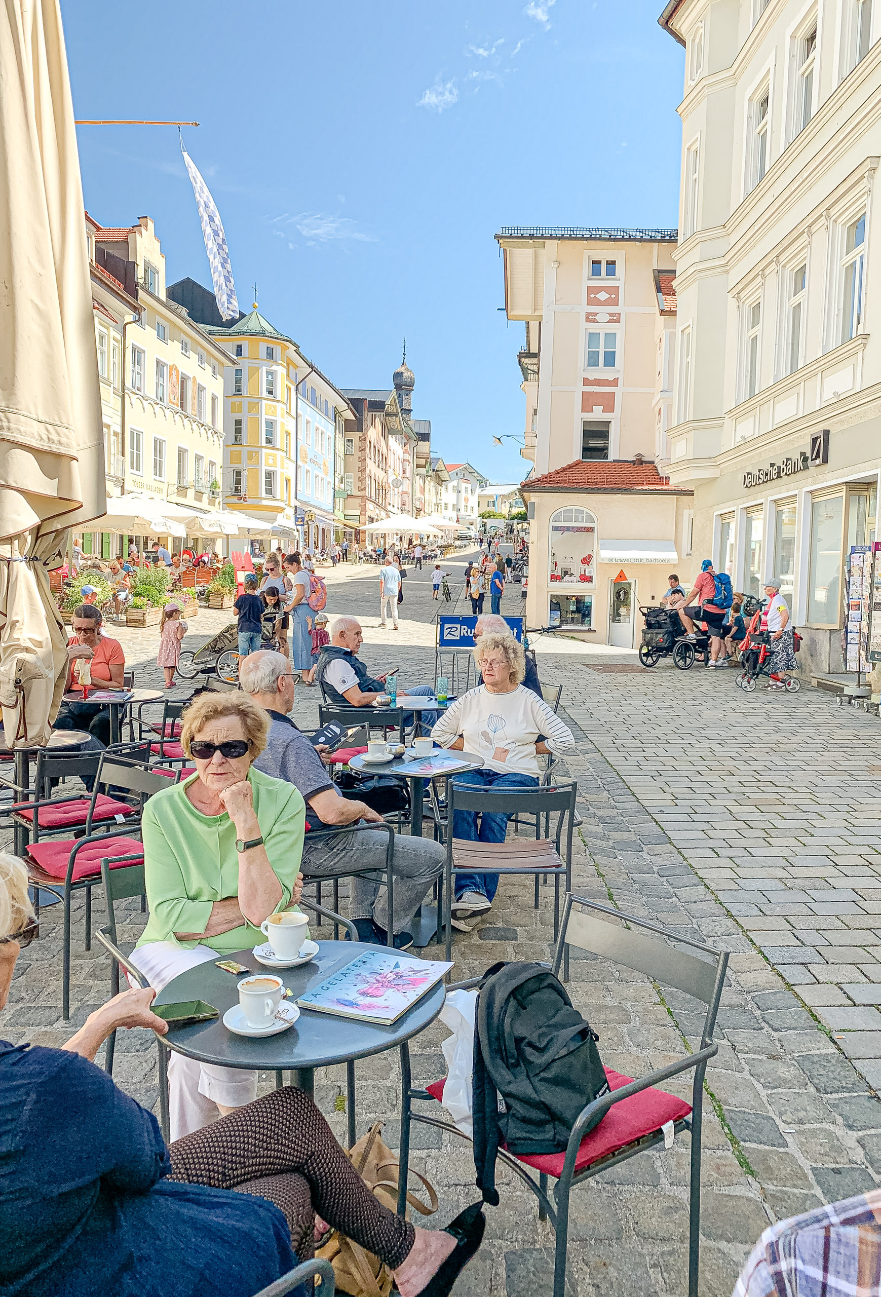 Menschen sitzen an Café-Tischen in einer sonnigen Fussgängergasse mit historischen Fassaden.
