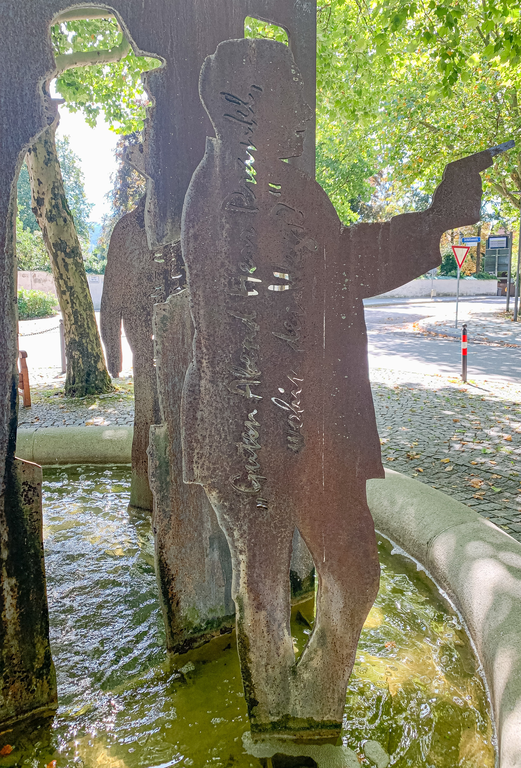 Rostige Metallsilhouetten von Männern mit Hüten stehen in einem Brunnen, eine Figur streckt einen Arm aus.