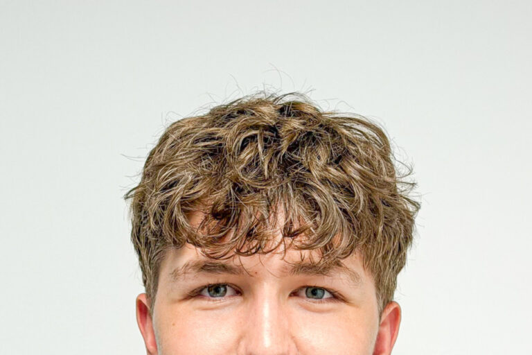 Junger Mann mit lockigem Haar und gestreiftem Poloshirt lächelt leicht in die Kamera.
