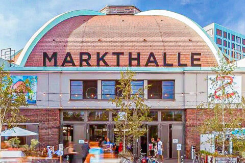 Historisches Backsteingebäude mit gewölbtem Dach und Schriftzug „Markthalle“, davor Passanten und Fahrräder.
