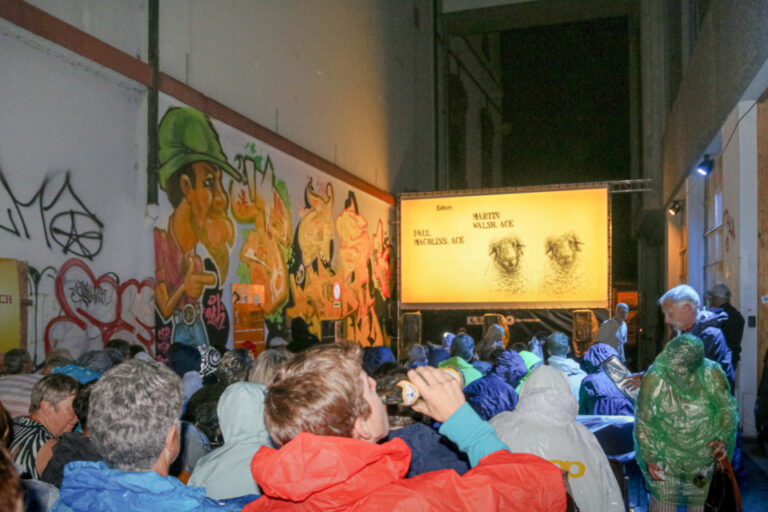 Menschen in Regenjacken sitzen nachts im Freien vor einer beleuchteten Kinoleinwand an einer bemalten Mauer.