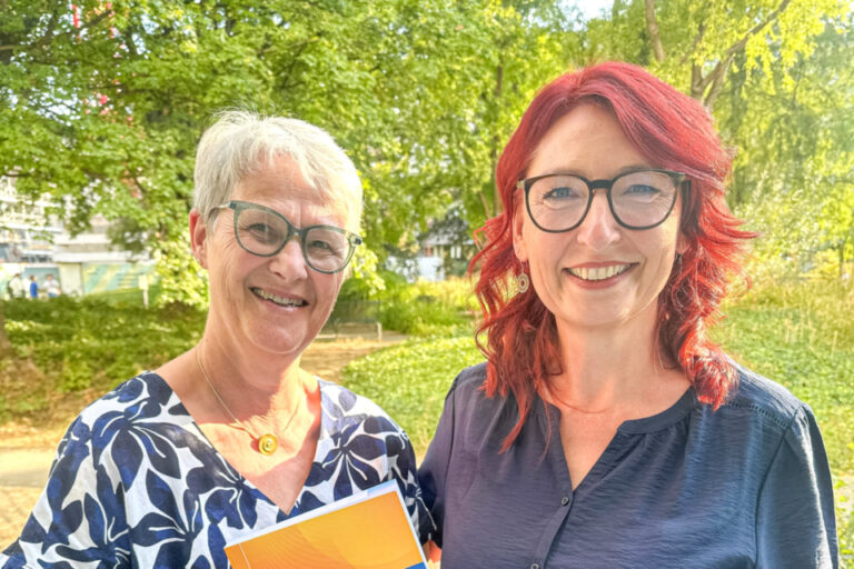 Zwei lächelnde Frauen stehen nebeneinander im Freien, eine hält ein Buch über Multiple Sklerose in den Händen.