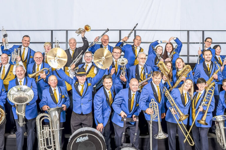 Grosse Musikkapelle in blauen Uniformjacken posiert mit Blasinstrumenten und Trommeln auf einer Bühne.