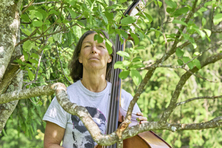 Eine Frau steht mit einem Kontrabass zwischen Bäumen auf einer grünen Wiese im Freien.