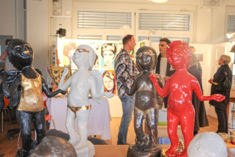 Bunte Skulpturen von Figuren im Vordergrund, im Hintergrund Besucher bei einer Kunstausstellung mit Gemälden.