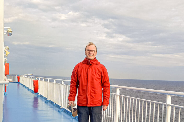 Ein Mann in roter Jacke steht auf dem blauen Deck eines Fährschiffs vor dem offenen Meer.