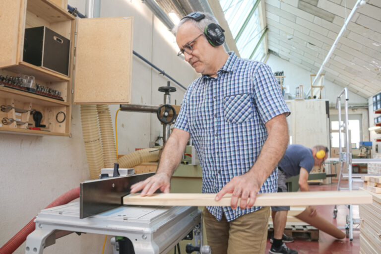Ein Mann mit Gehörschutz und Brille führt ein Holzbrett an einer Werkstattmaschine entlang.