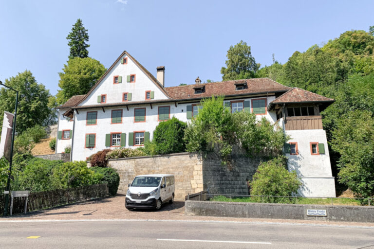 Ein grosses weisses Fachwerkhaus mit rotem Ziegeldach und grünen Fensterläden, umgeben von Bäumen, davor parkt ein Kleinbus.