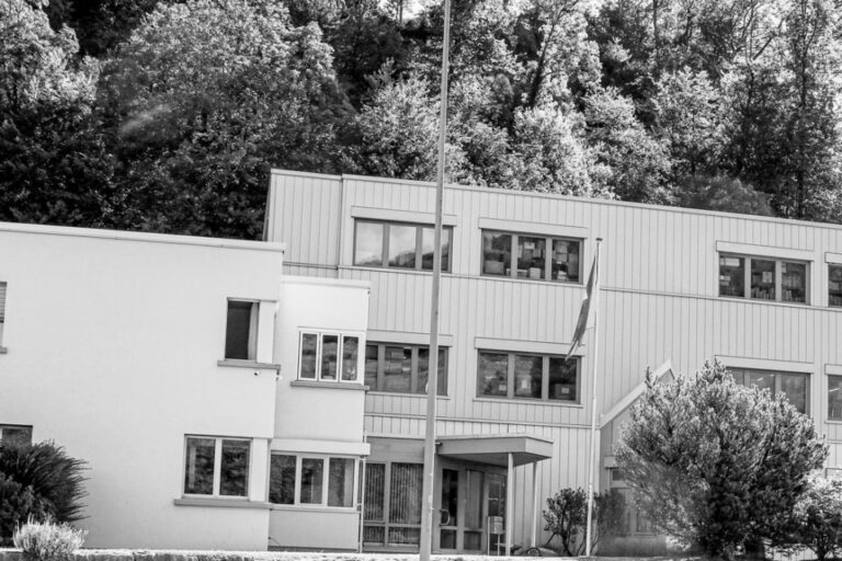 Ein modernes Firmengebäude mit Flachdach vor bewaldetem Hügel, Schwarz-Weiss-Aufnahme.