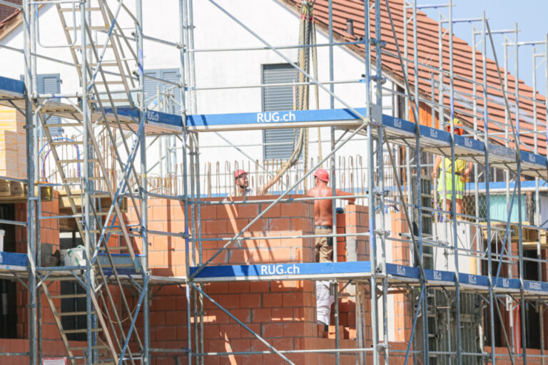 Bauarbeiter arbeiten auf Gerüsten an einem Rohbau mit Backsteinmauerwerk und Ziegeldach.