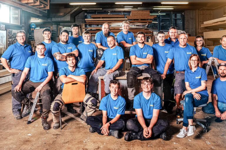 Gruppenfoto eines Teams in blauen Polo-Shirts und Arbeitshosen in einer Werkhalle einer Schreinerei.