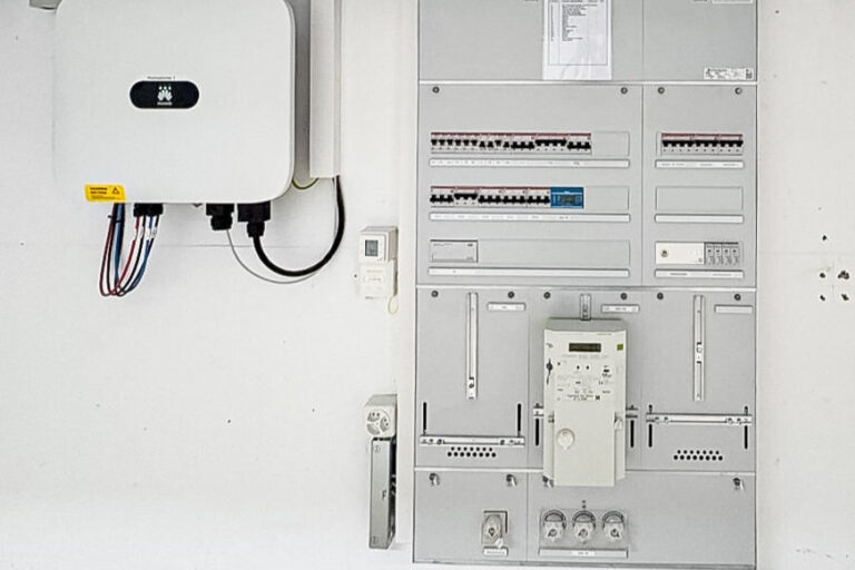 Elektrischer Schaltschrank mit Sicherungen und Zählern neben einem Wechselrichter an weisser Wand.