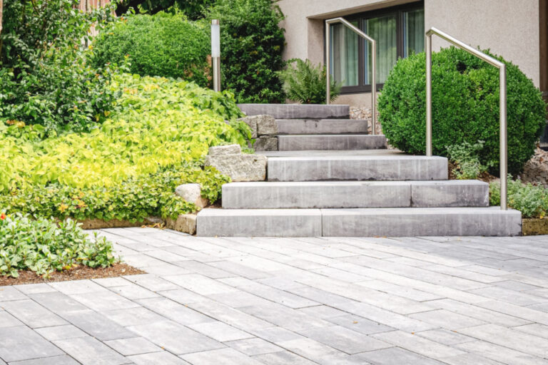 Gepflasterte Terrasse mit Steintreppe, Geländer und begrüntem Beet vor einem Hauseingang.