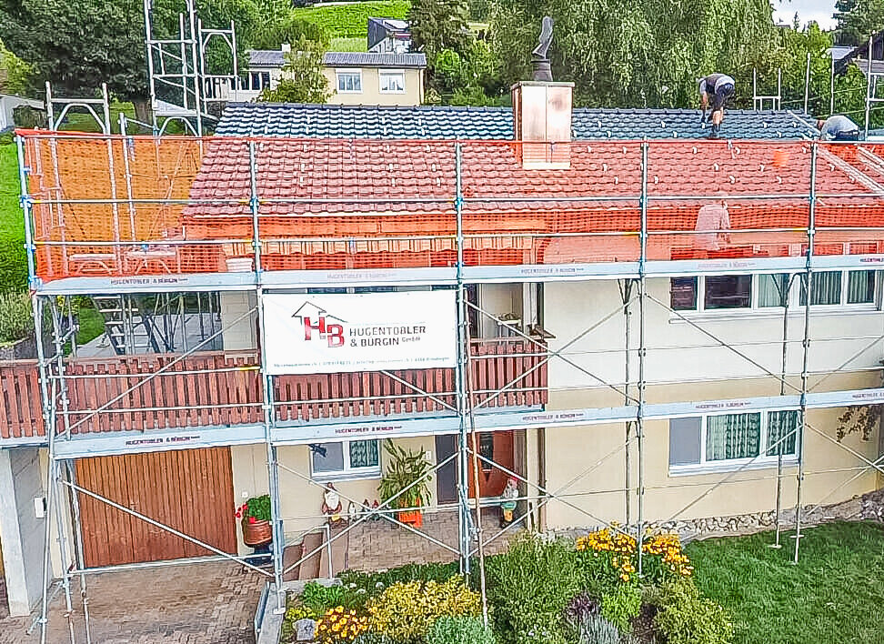 Ein Einfamilienhaus mit rotem Ziegeldach ist rundum mit Gerüst und orangem Schutznetz eingerüstet.