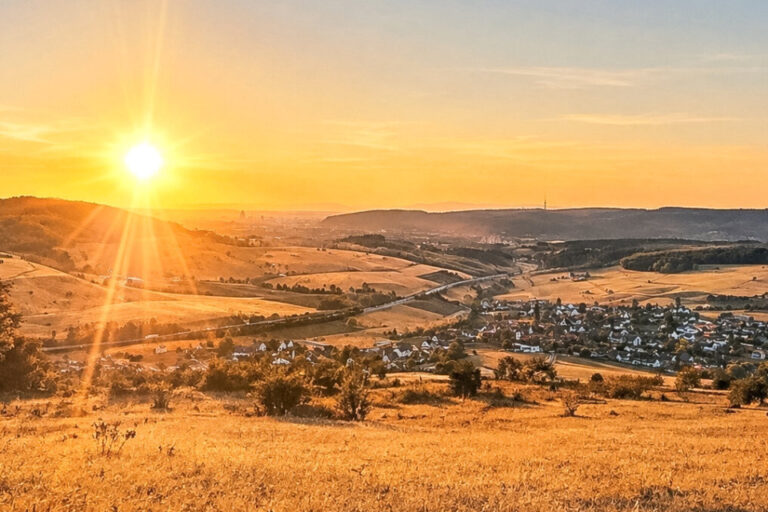 Sonnenuntergang über einem Tal mit Dorf, Feldern und Hügeln, im Vordergrund goldene Wiese.