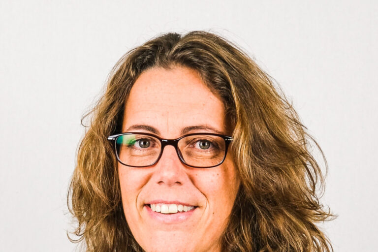 Frau mit lockigem, schulterlangem Haar und Brille lächelt in die Kamera, im dunklen Blazer.
