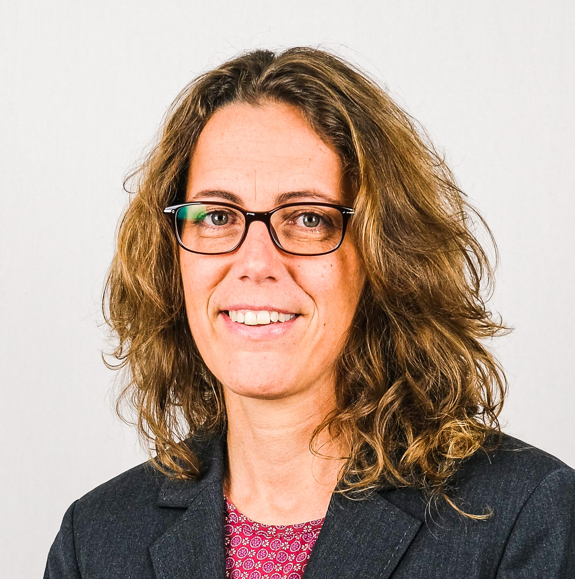Frau mit lockigem, schulterlangem Haar und Brille lächelt in die Kamera, im dunklen Blazer.
