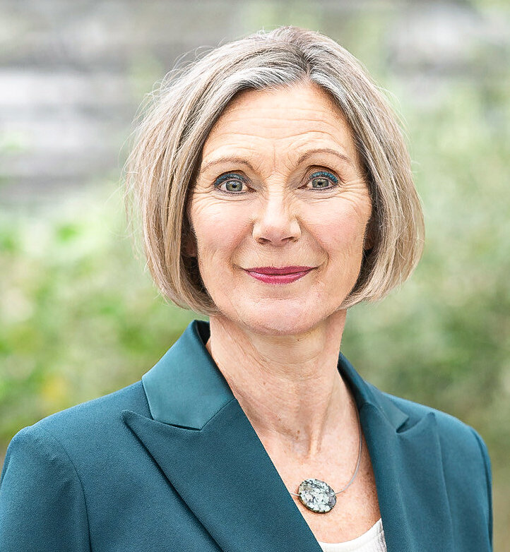Eine Frau mittleren Alters mit schulterlangem, grauem Haar in dunkelgrünem Blazer blickt lächelnd in die Kamera.