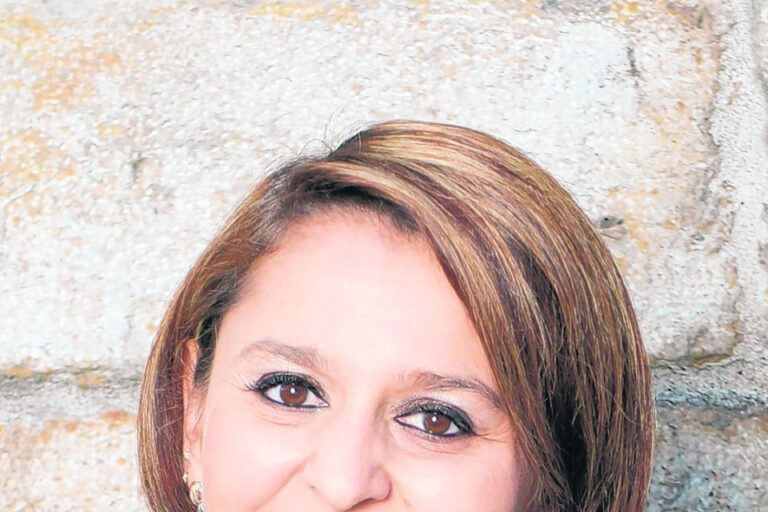 Eine lächelnde Frau mit schulterlangem braunem Haar trägt einen schwarzen Blazer vor einer Steinwand.