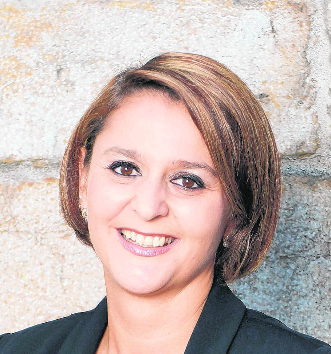 Eine lächelnde Frau mit schulterlangem braunem Haar trägt einen schwarzen Blazer vor einer Steinwand.