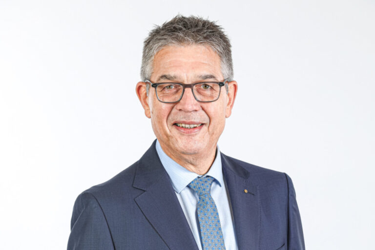 Ein älterer Mann mit Brille und grauem Haar im dunkelblauen Anzug lächelt vor hellem Hintergrund.