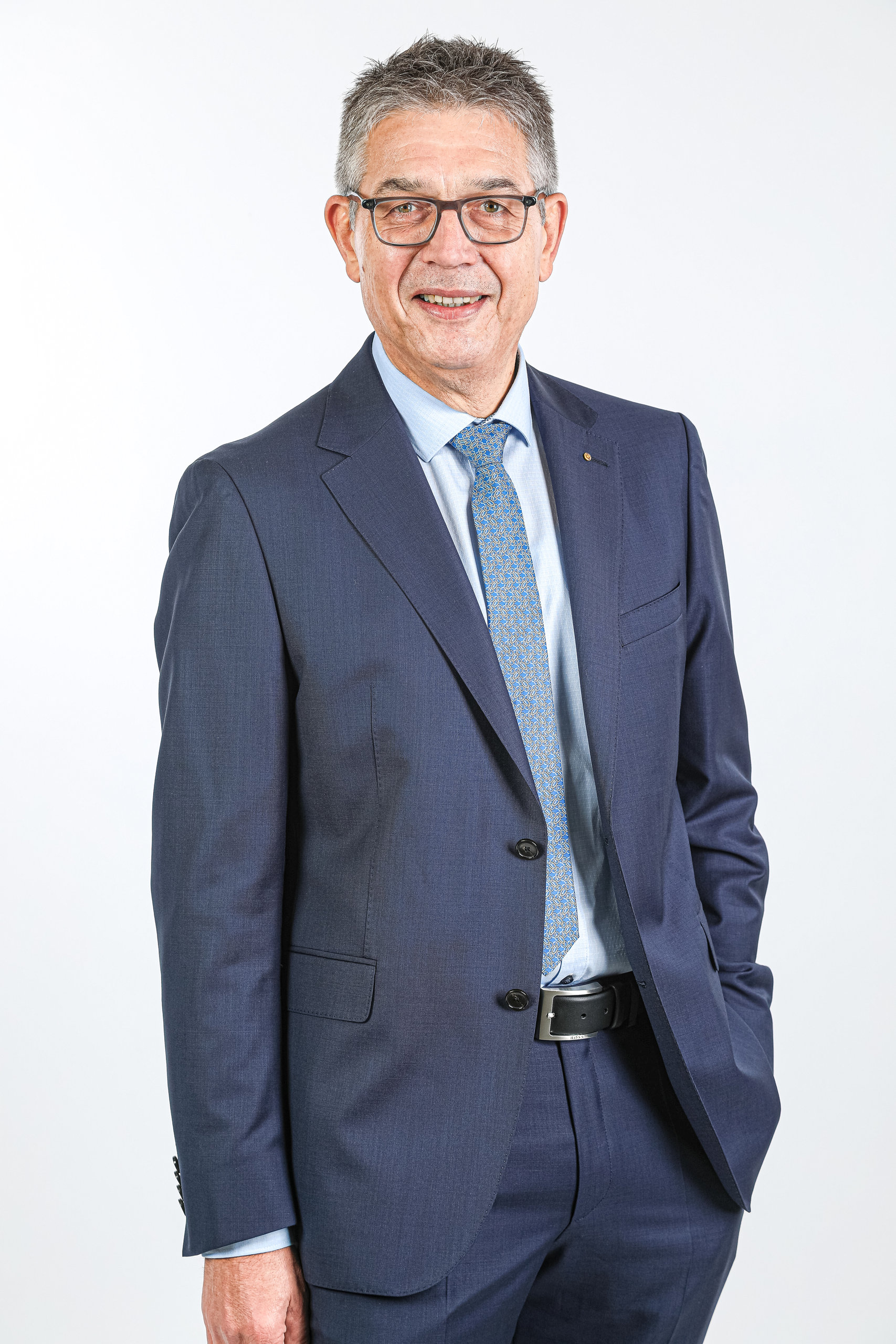 Ein älterer Mann mit Brille und grauem Haar im dunkelblauen Anzug lächelt vor hellem Hintergrund.