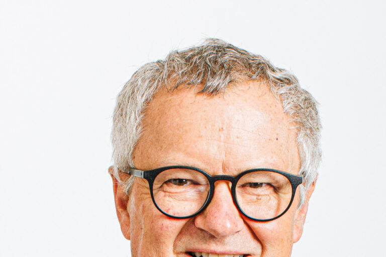 Ein älterer Mann mit grauen Haaren, Brille und Anzug lächelt in die Kamera.