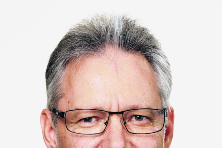 Ein älterer Mann mit grauem Haar, Brille und Bart trägt Anzug mit Krawatte und lächelt.