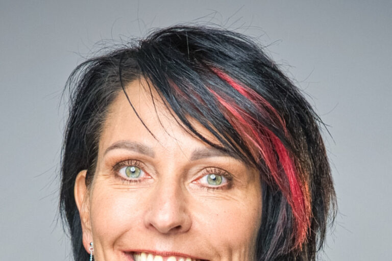 Lächelnde Frau mit schwarzen, rot gesträhnten Haaren in weissem Rollkragenpullover und dunklem Blazer.