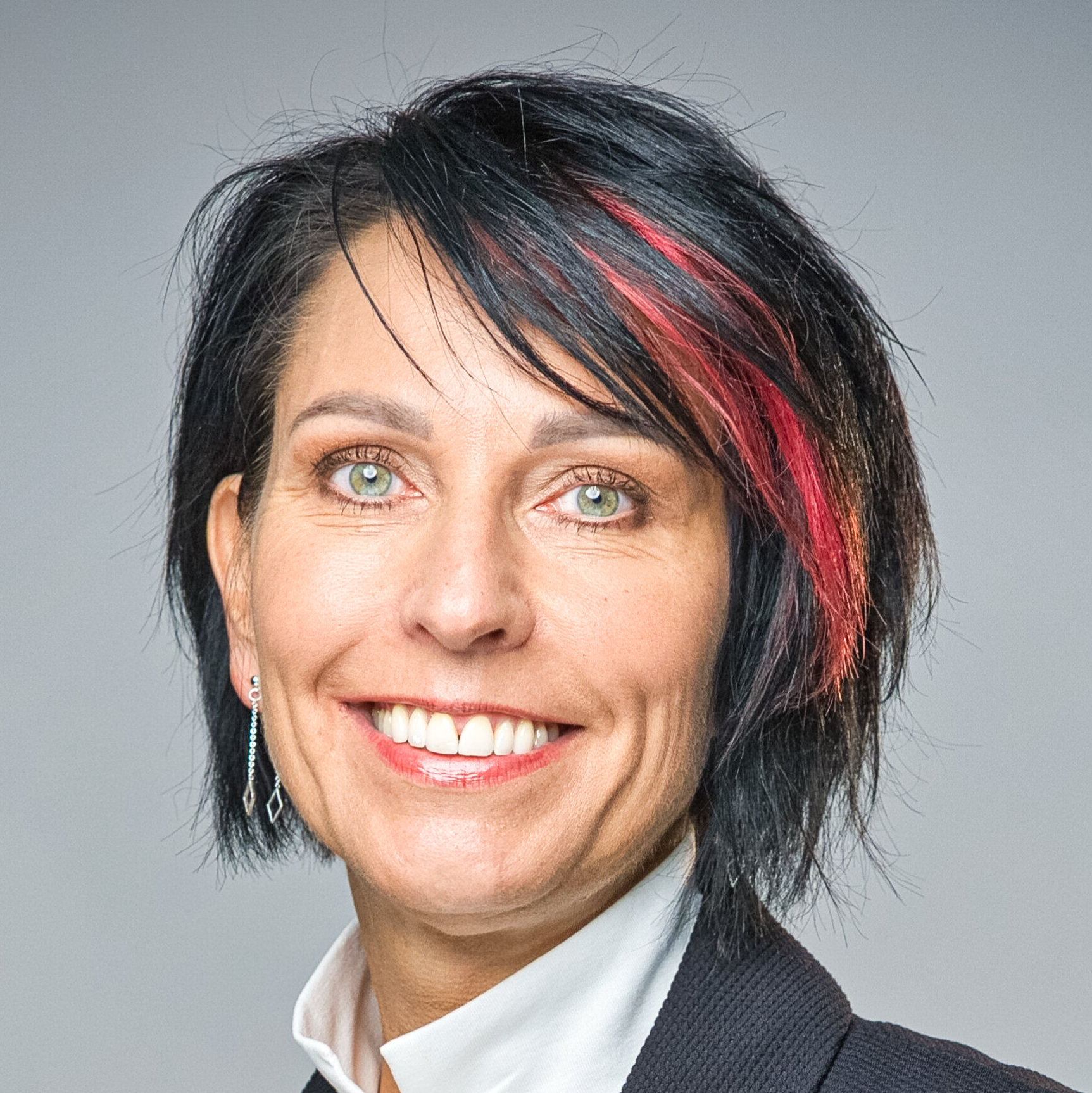 Lächelnde Frau mit schwarzen, rot gesträhnten Haaren in weissem Rollkragenpullover und dunklem Blazer.