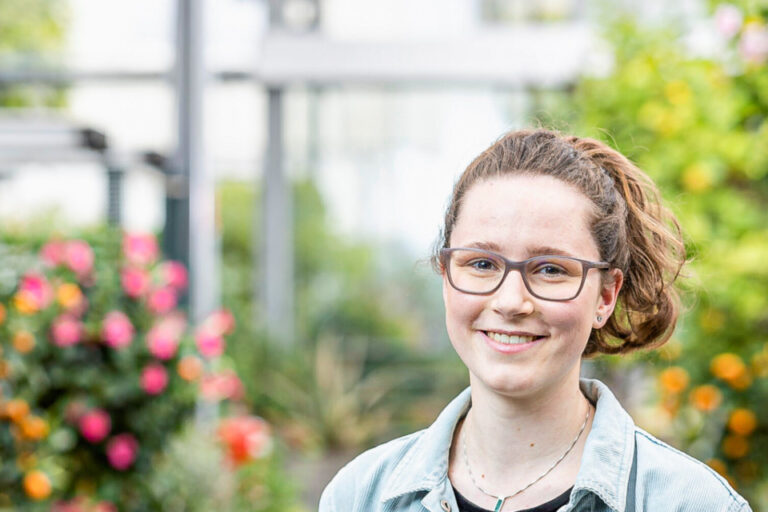 Eine junge Frau mit Brille und Jeansjacke lächelt vor blühenden Pflanzen in einem Gewächshaus.