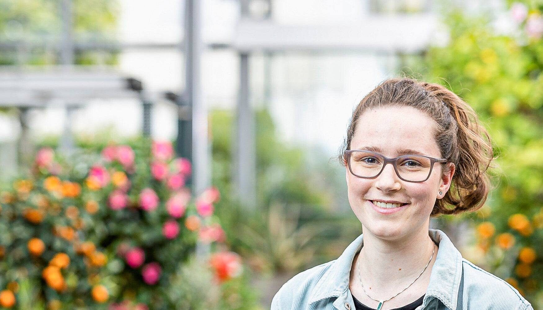 Eine junge Frau mit Brille und Jeansjacke lächelt vor blühenden Pflanzen in einem Gewächshaus.