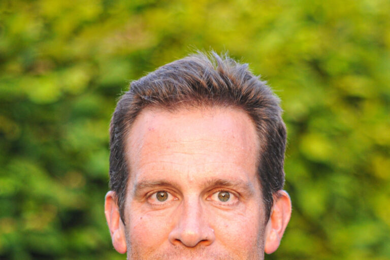 Ein Mann mittleren Alters mit kurzen braunen Haaren im Karohemd lächelt vor grünem Hintergrund.