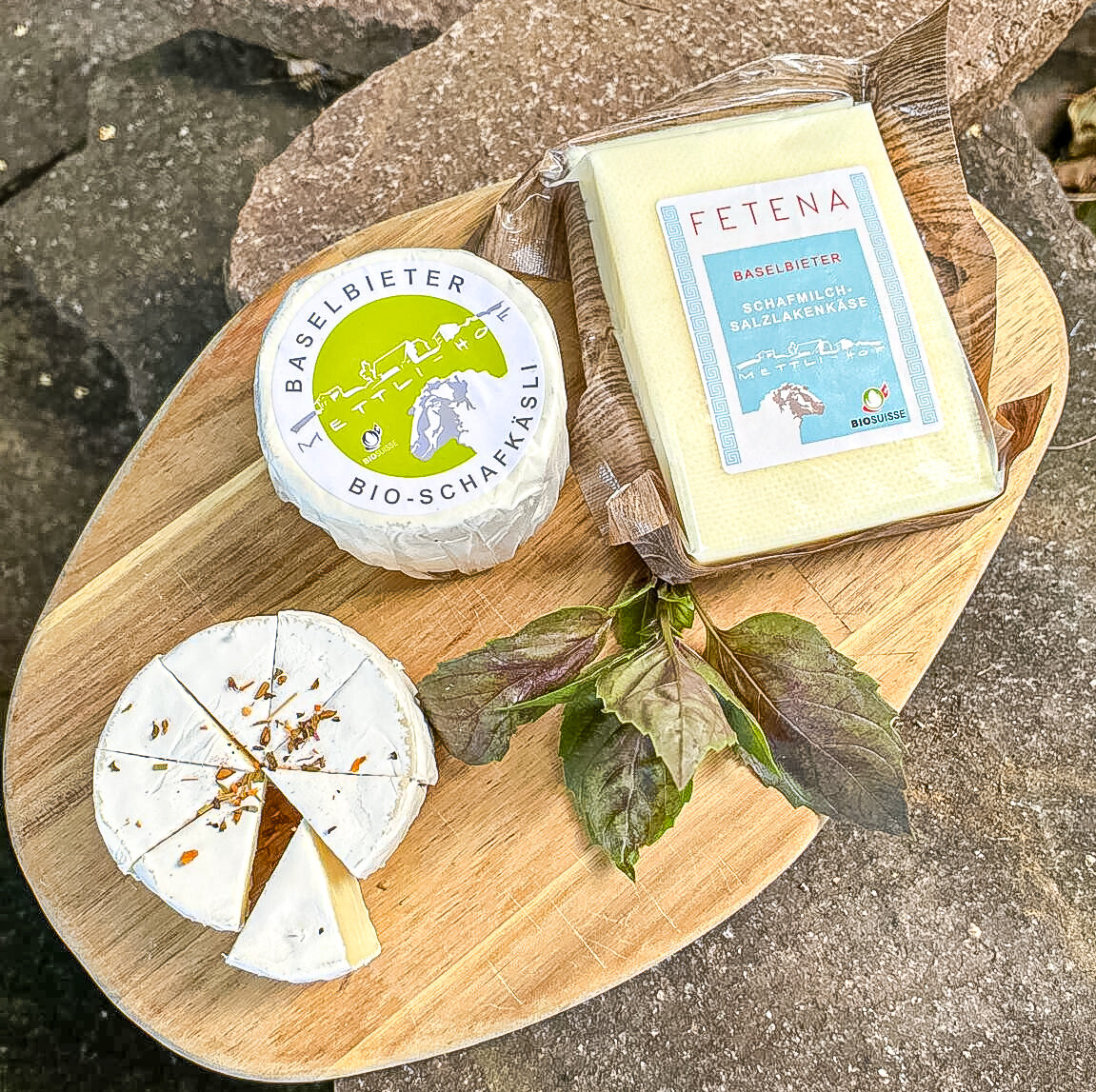 Käseplatte mit Schafkäse, verpacktem Käsestück und frischen Kräutern auf Holzbrett und Steinen.