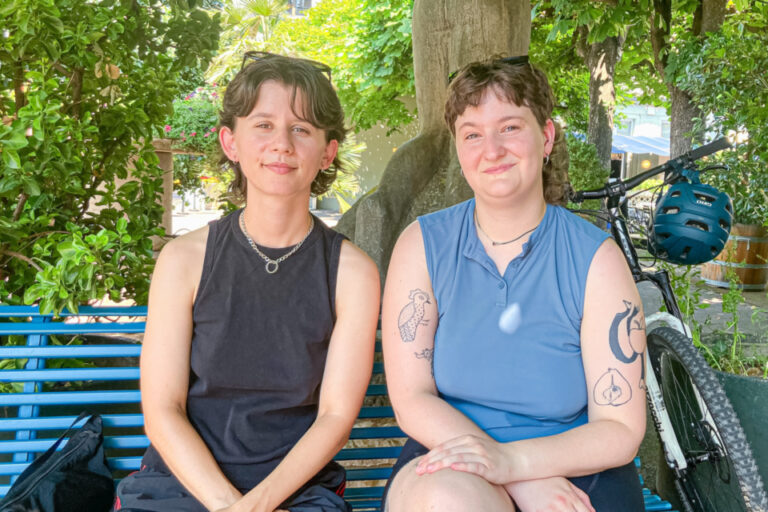 Zwei lächelnde Frauen sitzen nebeneinander auf einer blauen Bank unter einem Baum, daneben ein Fahrrad.