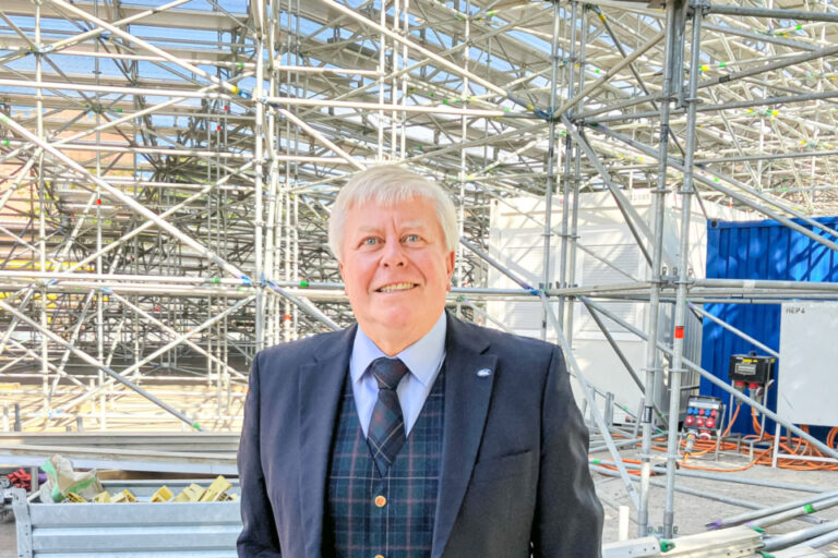 Ein lächelnder Mann im Anzug mit Karo-Weste steht vor einer Baustelle mit Gerüsten.