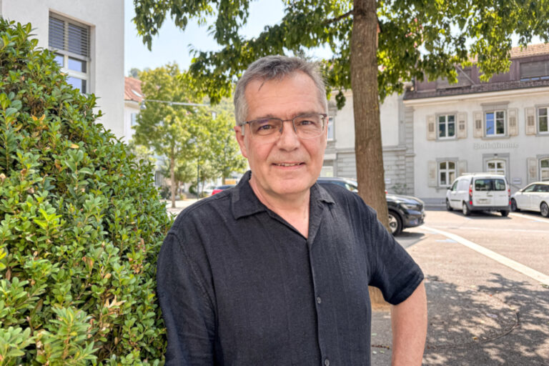 Ein Mann mit Brille und grauem Haar in dunklem Hemd steht lächelnd vor einer Hecke auf einem Platz.