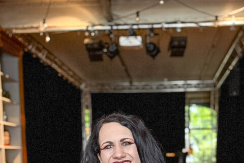 Eine lachende Frau mit schwarzen Haaren und roten Dreadlock-Strähnen sitzt an einem Tisch in einem Veranstaltungsraum.