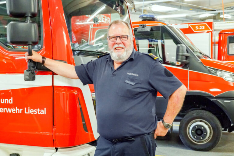 Ein lächelnder Mann mit Brille und Bart steht an ein Feuerwehrfahrzeug gelehnt in einer Fahrzeughalle.
