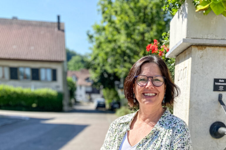 Lächelnde Frau mittleren Alters sitzt an einem steinernen Dorfbrunnen mit Blumen in der Sonne.