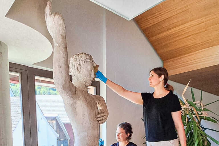 Zwei Frauen mit blauen Handschuhen reinigen eine lebensgrosse Steinstatue eines nackten Mannes in einem Raum.