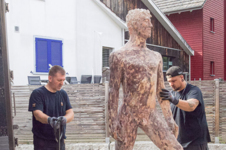 Zwei Männer stellen eine grosse menschenförmige Skulptur aus Metall vor einem Lastwagen auf.