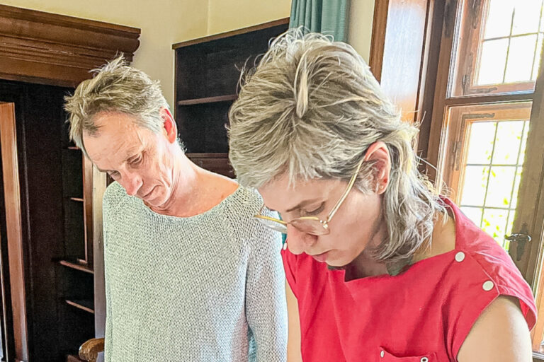 Ein Mann und eine Frau beugen sich über einen Tisch mit Karten und Zeichnungen in einem Zimmer mit Holzvertäfelung.