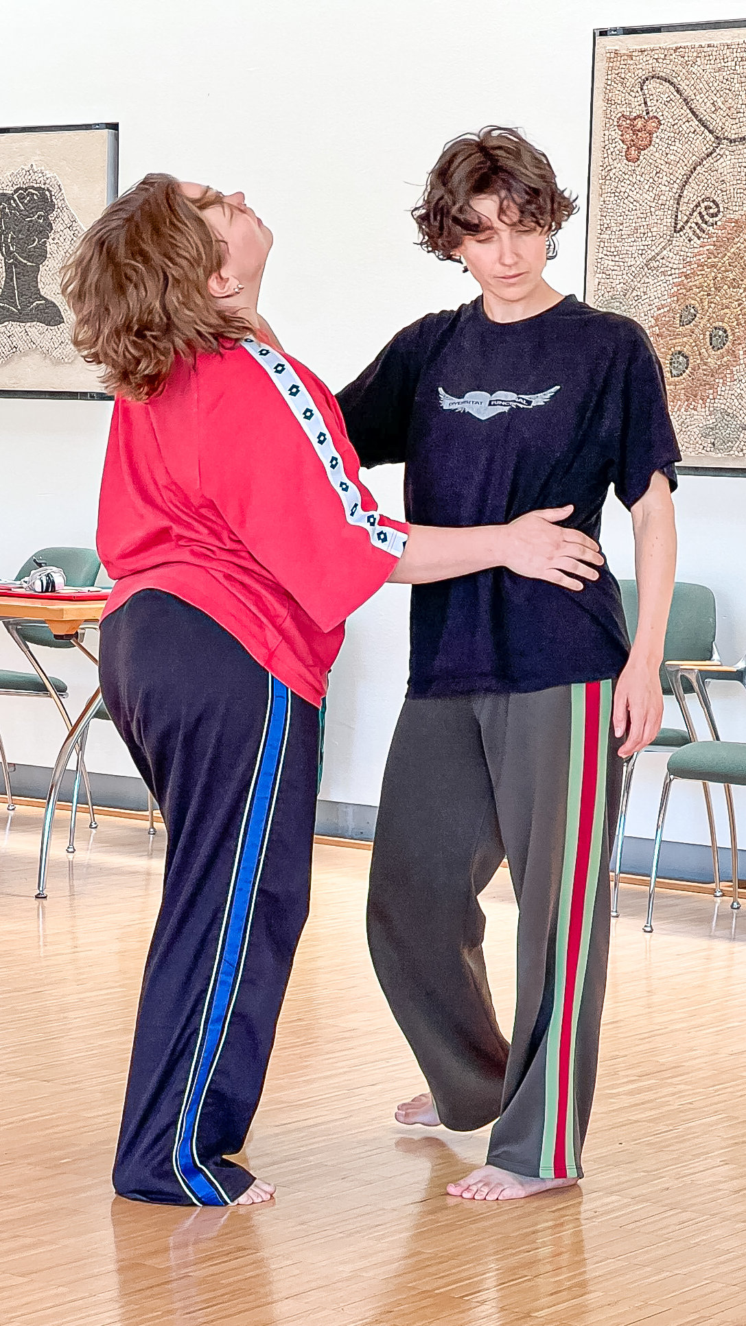 Zwei Frauen stehen sich barfuss in einem Saal gegenüber, eine legt die Hand an die Hüfte der anderen.