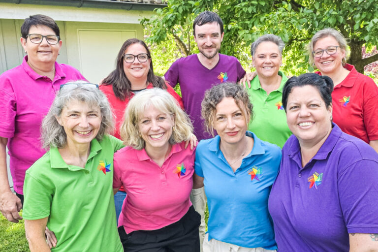 Acht lächelnde Personen in bunten Poloshirts stehen und knien im Garten vor Bäumen.