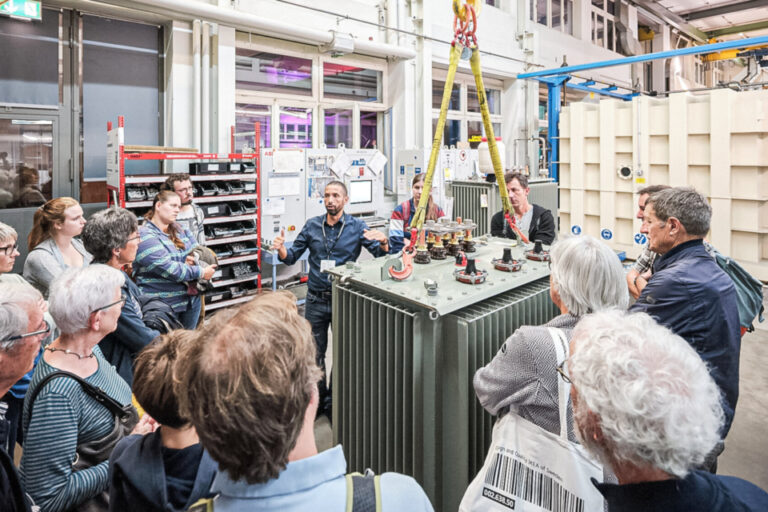 Besuchergruppe steht um einen grossen Transformator in einer Werkshalle und hört einem Mitarbeiter zu.