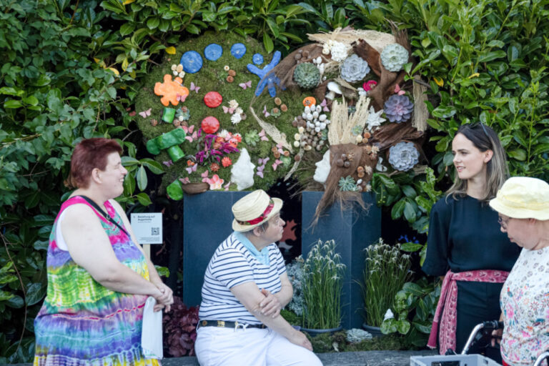 Menschen unterhalten sich vor einer mit bunten Blumen und Naturmaterialien geschmückten Hecke.