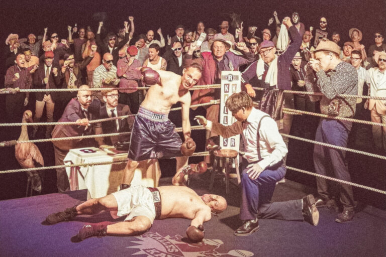 Ein Boxer steht triumphierend über einem am Boden liegenden Gegner im Ring, jubelnde Zuschauer im Hintergrund.