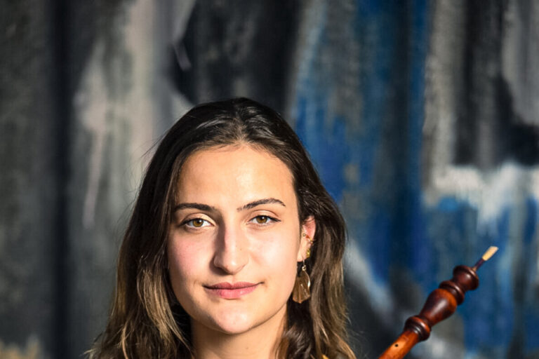 Junge Frau mit lockigem Haar hält ein Holzblasinstrument vor gemusterter Bluse, Graffiti-Wand im Hintergrund.